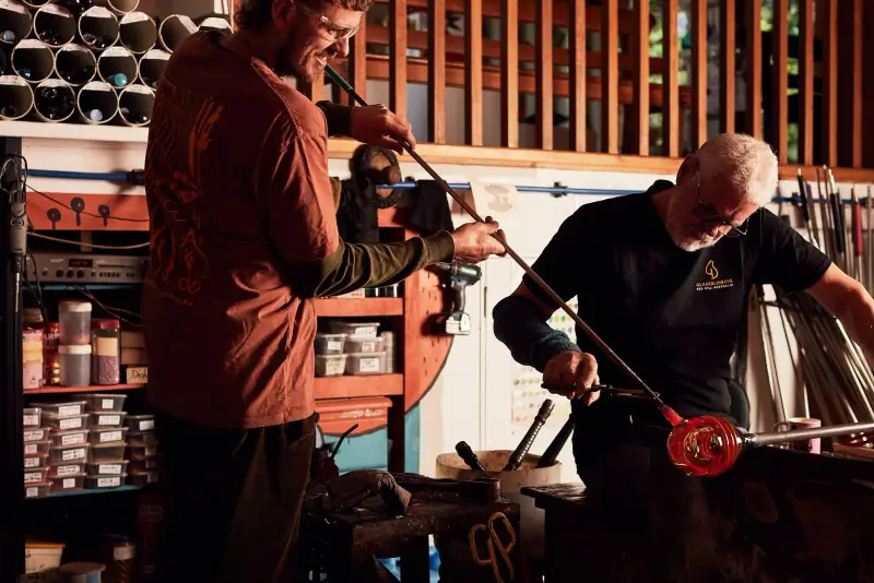 Two glassblowers turning a glowing piece of glass on a blowpipe at the bench in the Gordon Studio hot shop