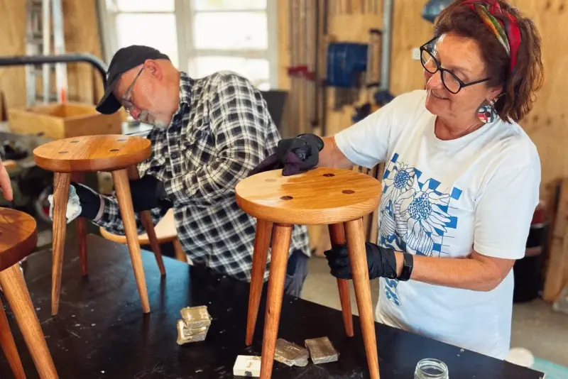 Two people on a course sanding their stools at a workbench in the timber-lined workshop
