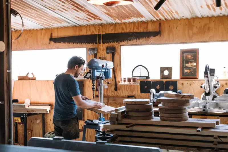 A woodworker at the drill press in the timber-lined workshop, with round stool seats stacked on the bench