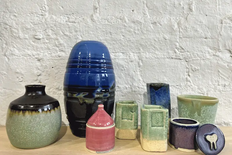 Glazed vases, cups and a lidded jar lined up against a white brick wall