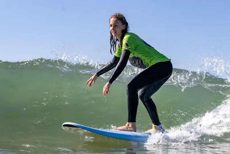 A surfer in a green rash vest standing up on a soft board in a small wave