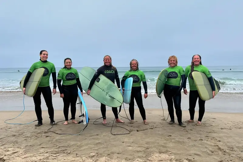 A women's surf group in wetsuits and green rash vests standing on the beach with their boards