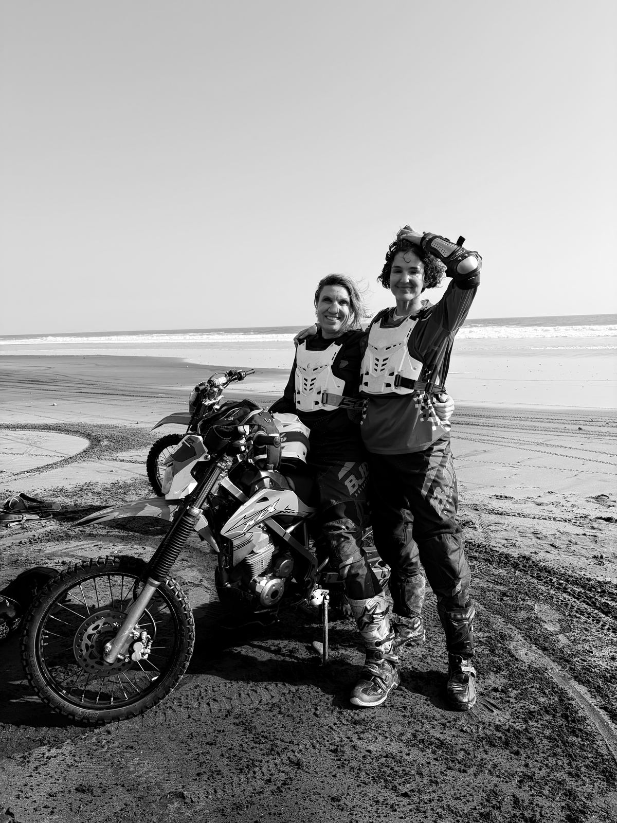 Simone, owner of Salty Cowboy, and Ro, RealDays founder, in motocross gear beside a dirt bike on a black-sand beach in Bali